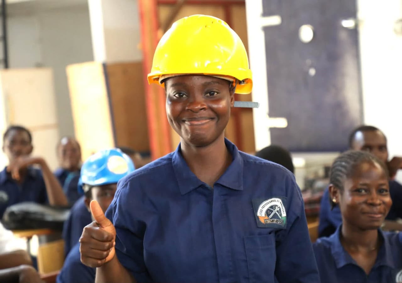 Élève souriante en combinaison bleue et casque jaune, pouce levé, dans l'atelier
